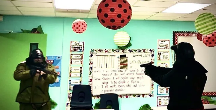Two role players in protective gear and helmets face each other with training weapons in an elementary classroom during a force-on-force scenario